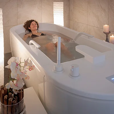 Frau entspannt in moderner Badewanne mit Wasser, Kerzen, Orchidee und sanfter Beleuchtung.