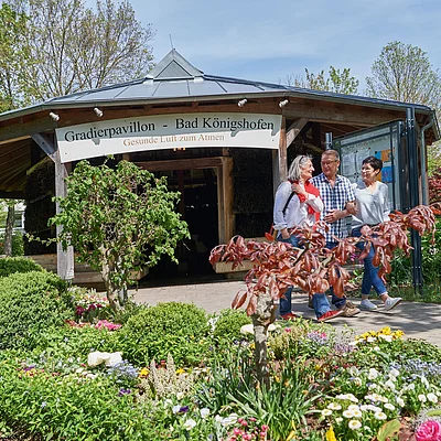 Drei Personen gehen vor dem Gradierpavillon Bad Königshofen durch einen Garten mit Blumen und Sträuchern.
