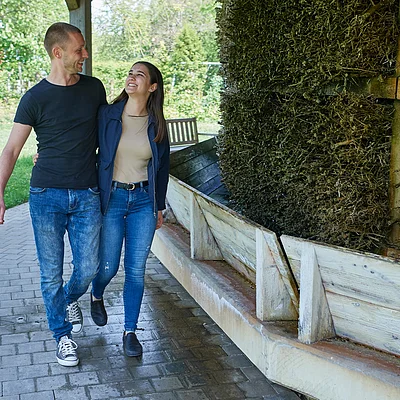 Paar geht Arm in Arm auf gepflastertem Weg neben einer großen Holzkonstruktion mit Reisig.