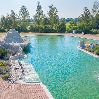 Außenpool mit klarem Wasser, Felsenstruktur und vier Personen am Beckenrand, umgeben von Bäumen und Wiese.