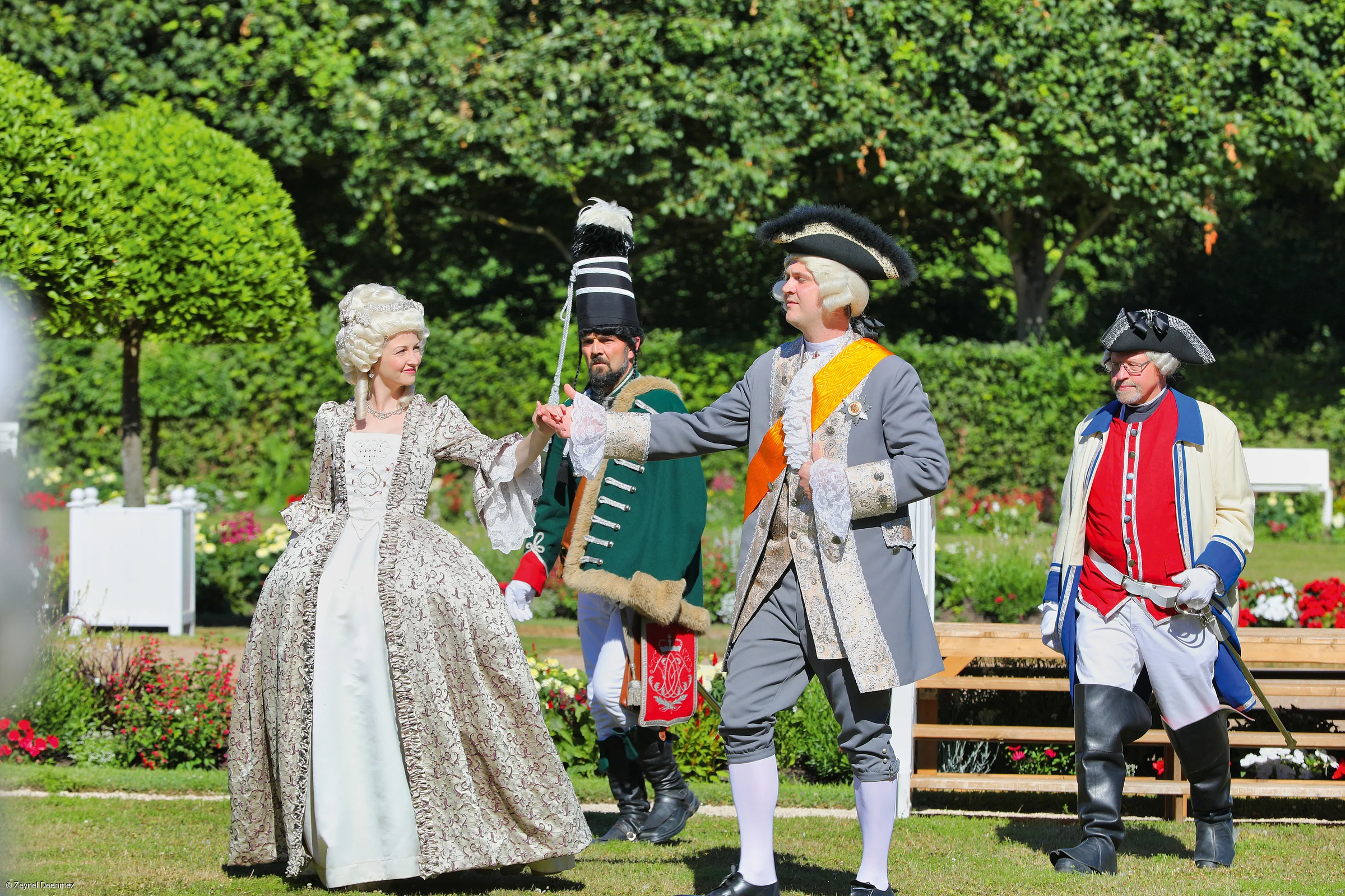 Vier Personen in historischen Kostümen aus dem Rokoko im Garten, zwei tanzen Händchen haltend.