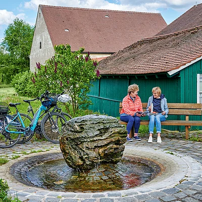 Zwei Frauen sitzen auf einer Bank neben einem Brunnen mit großem Stein, Fahrräder stehen daneben, grüne Holzhütte im Hintergrund.