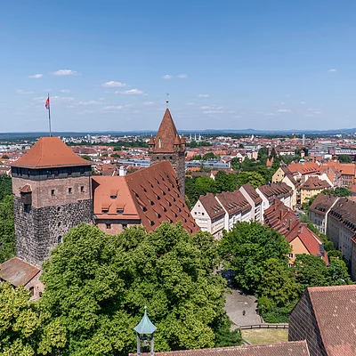 Blick über eine Stadt mit historischen Gebäuden, roten Dächern und vielen Bäumen unter blauem Himmel.