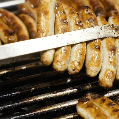 Mehrere Bratwürste werden mit einer Metallzange auf einem Grill gewendet.