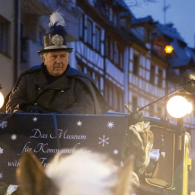 Mann in Postuniform fährt abends in geschmückter Kutsche durch eine Altstadtstraße mit Fachwerkhäusern.