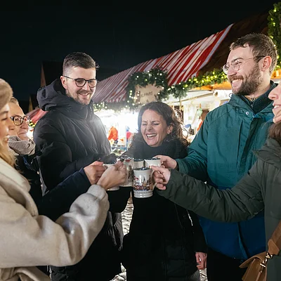 Sechs Personen stoßen mit Tassen bei einem beleuchteten Weihnachtsmarktstand bei Nacht an.