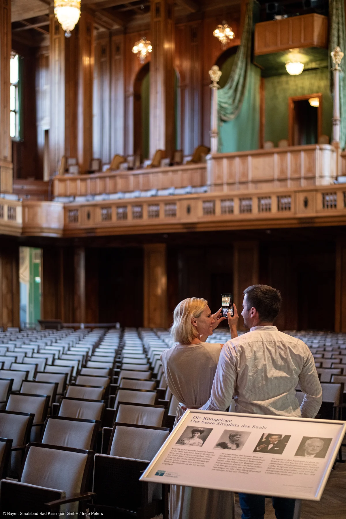 Zwei Personen fotografieren in einem großen Saal mit vielen Sitzreihen und einer hölzernen Königsloge.