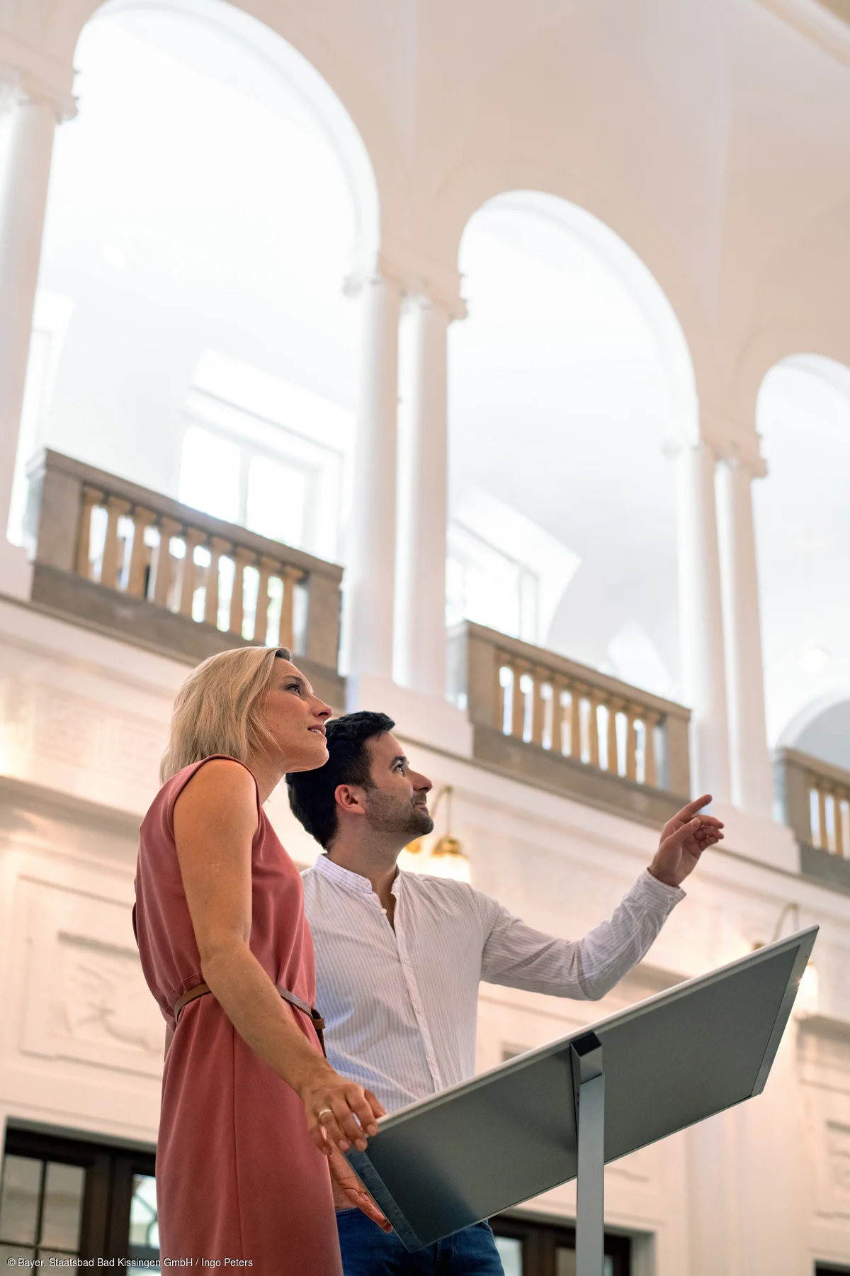 Zwei Personen betrachten und zeigen auf eine Ausstellungstafel in einem hellen Gebäude mit hohen Bögen.