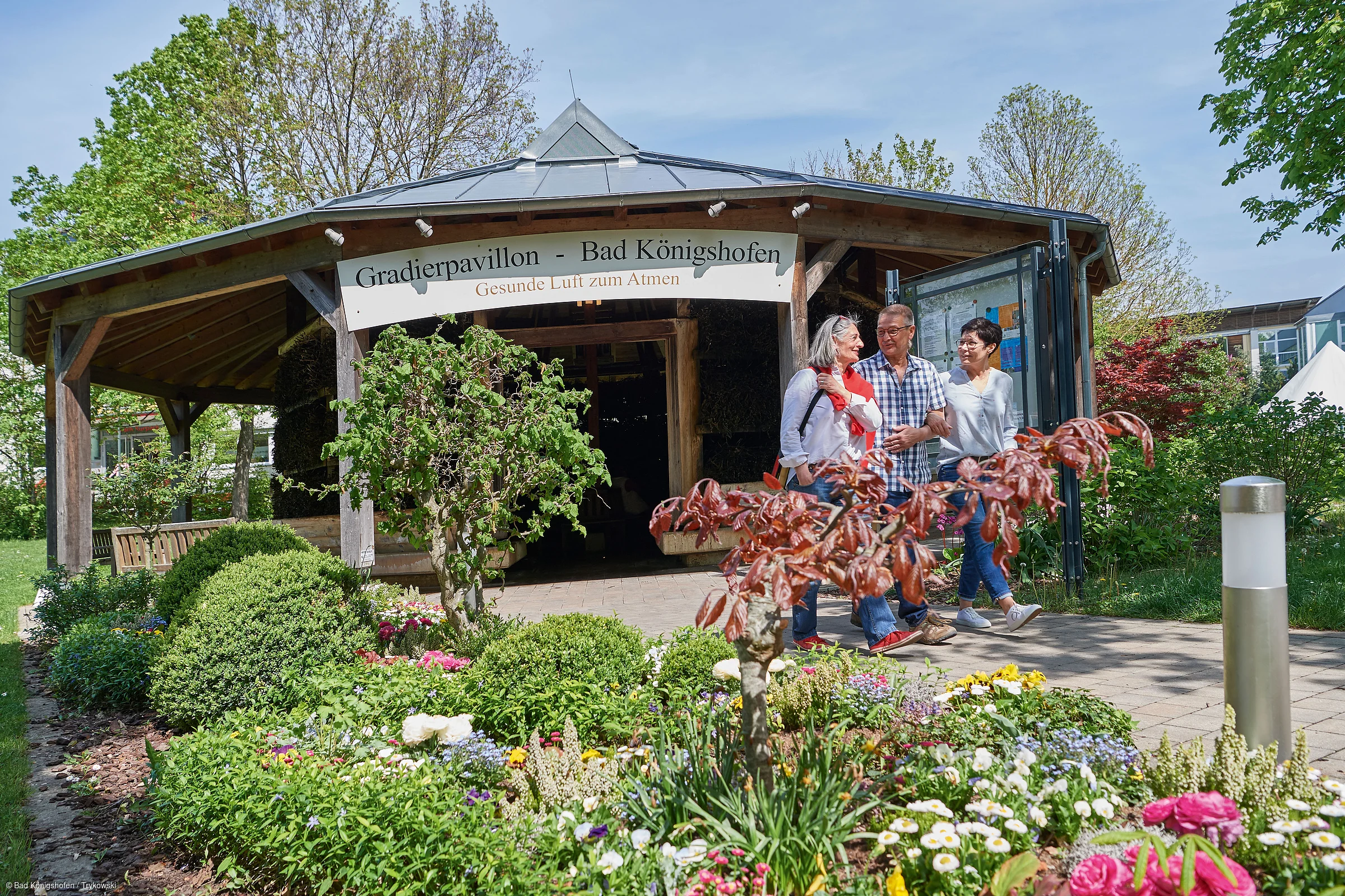 Drei Personen gehen vor dem Gradierpavillon Bad Königshofen durch einen Garten mit Blumen und Sträuchern.