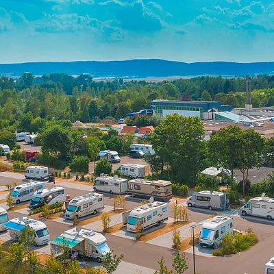 Luftaufnahme eines Campingplatzes mit Wohnmobilen, Bäumen und Gebäuden im Hintergrund bei klarem Himmel.