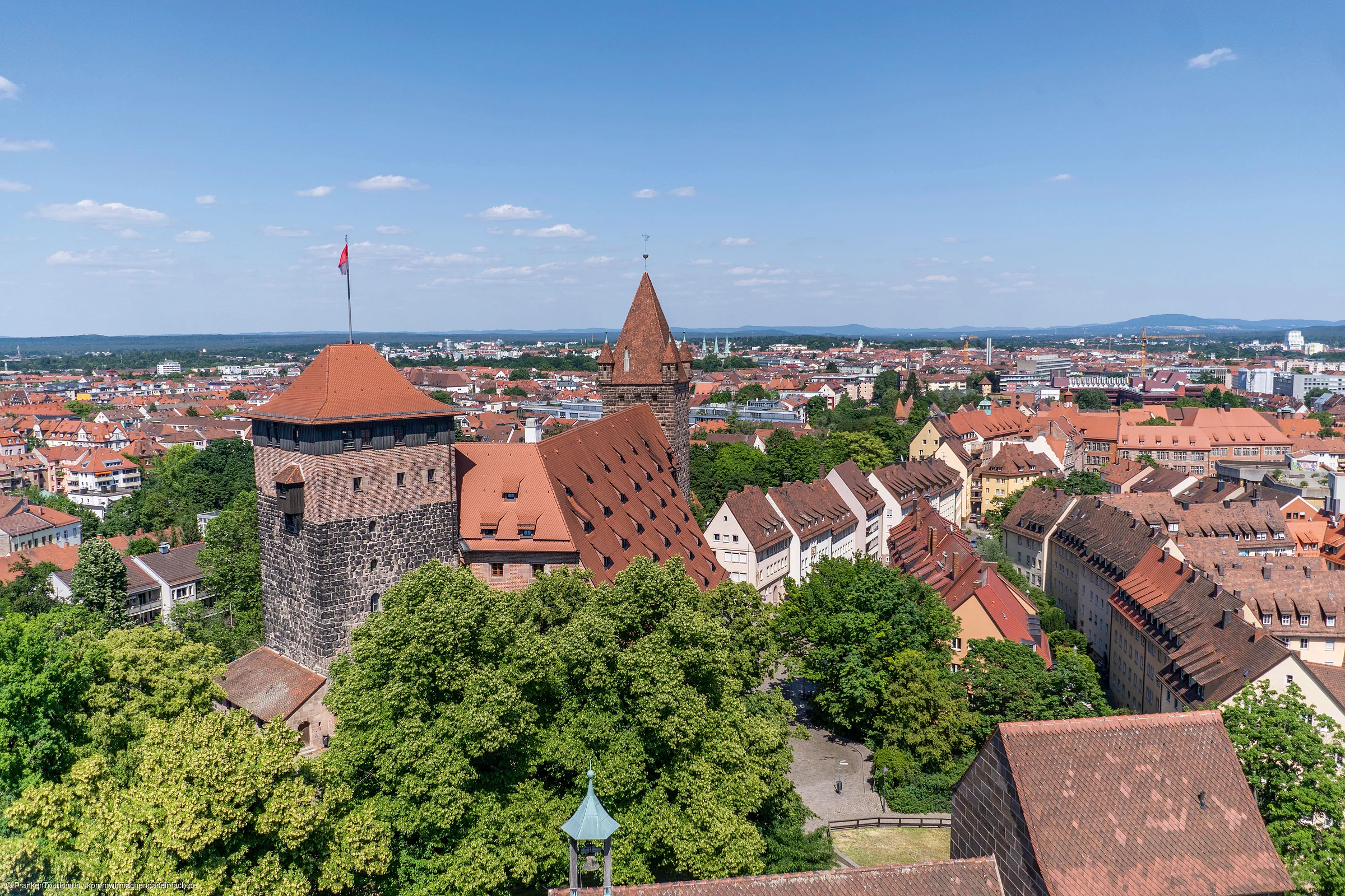 Blick über eine Stadt mit historischen Gebäuden, roten Dächern und vielen Bäumen unter blauem Himmel.