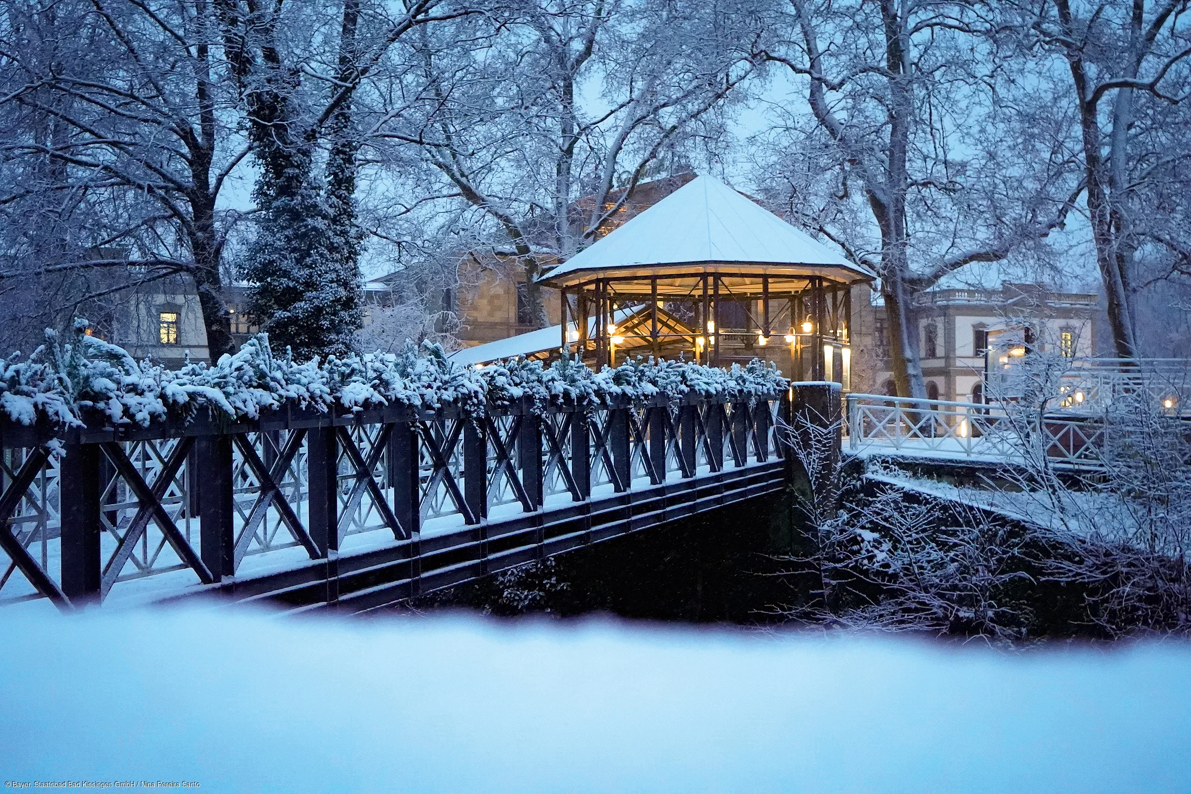 Schneebedeckte Brücke mit Lichterzelt und Bäumen in winterlicher Abenddämmerung.