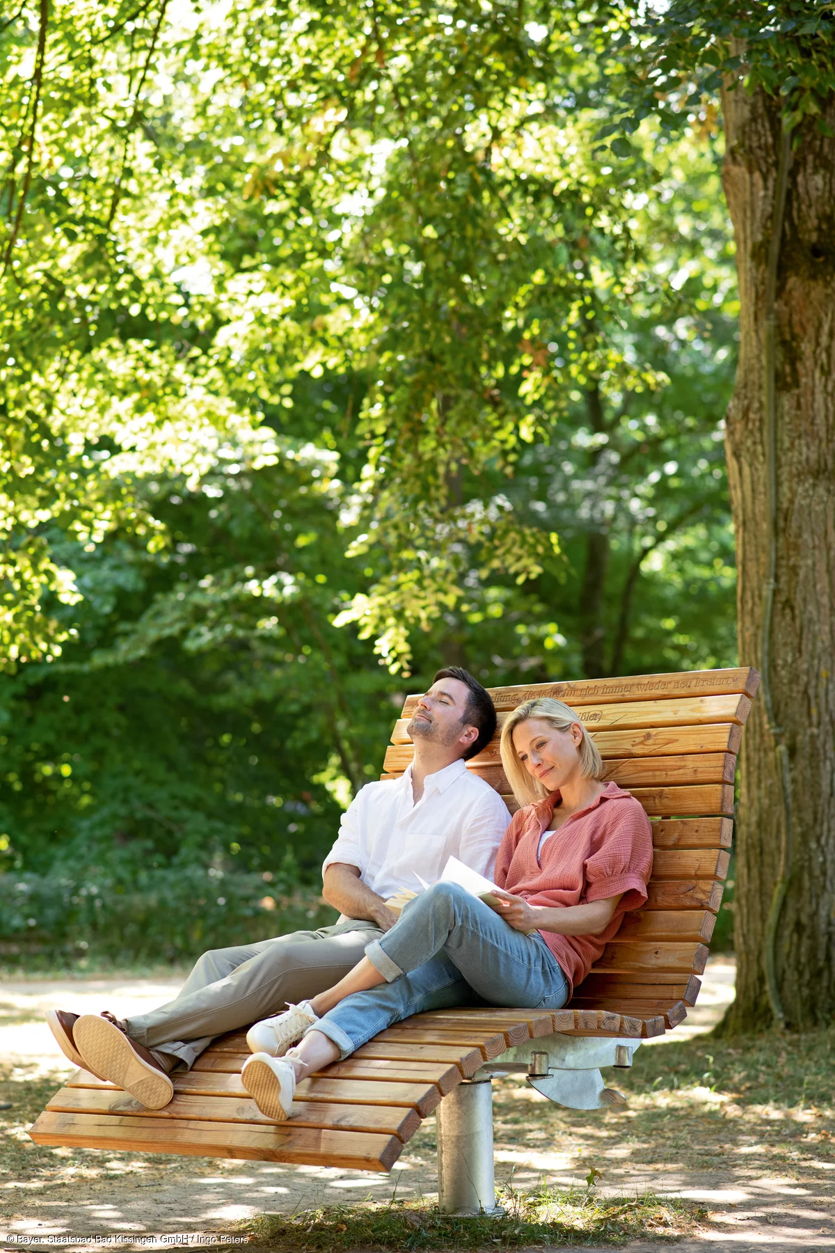 Paar sitzt entspannt auf geschwungener Holzbank im Park unter Bäumen bei Sonnenschein