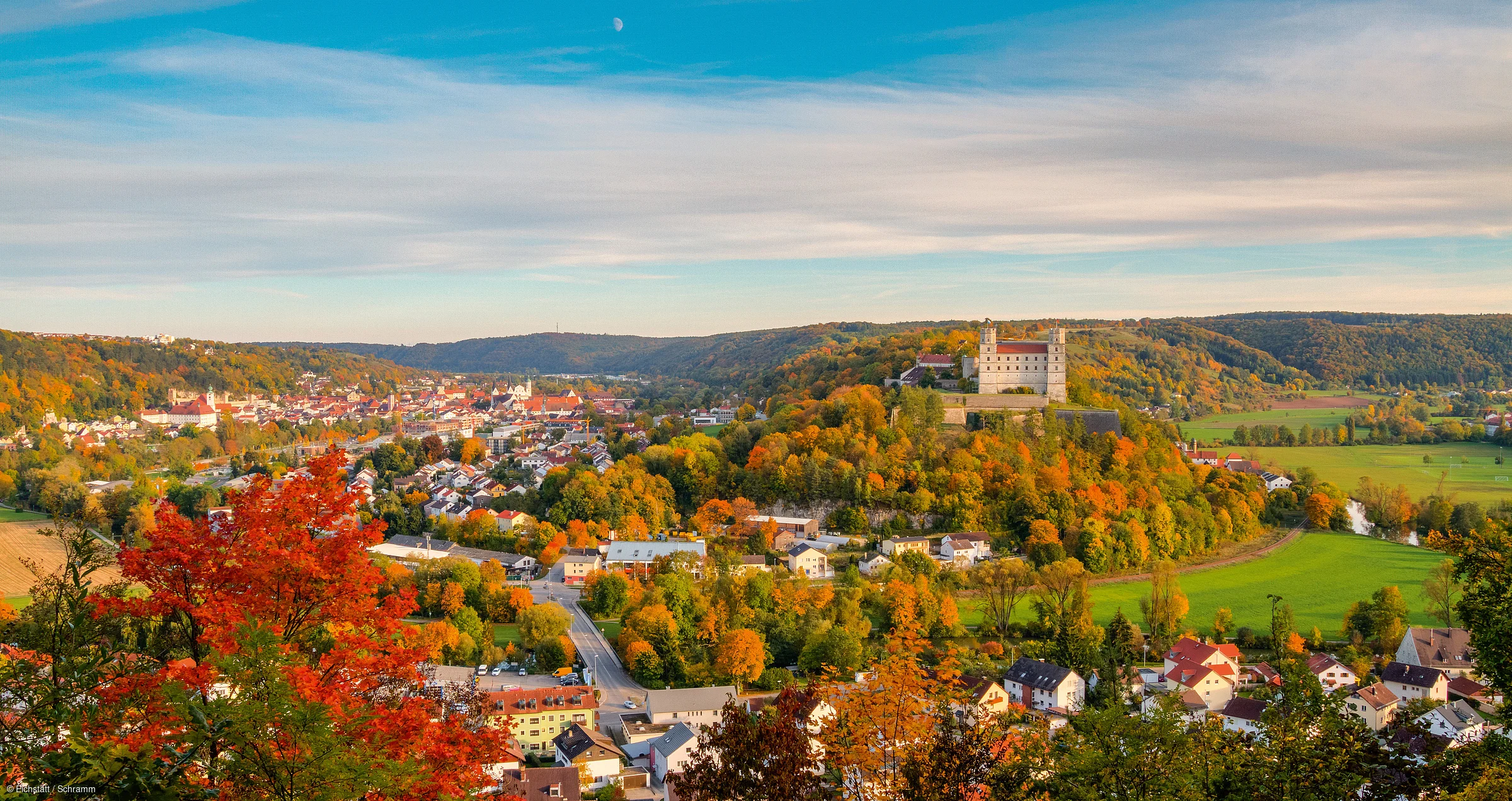 Herbstliche Landschaft mit Schloss auf Hügel, bunten Bäumen und Dorf unter blauem Himmel.