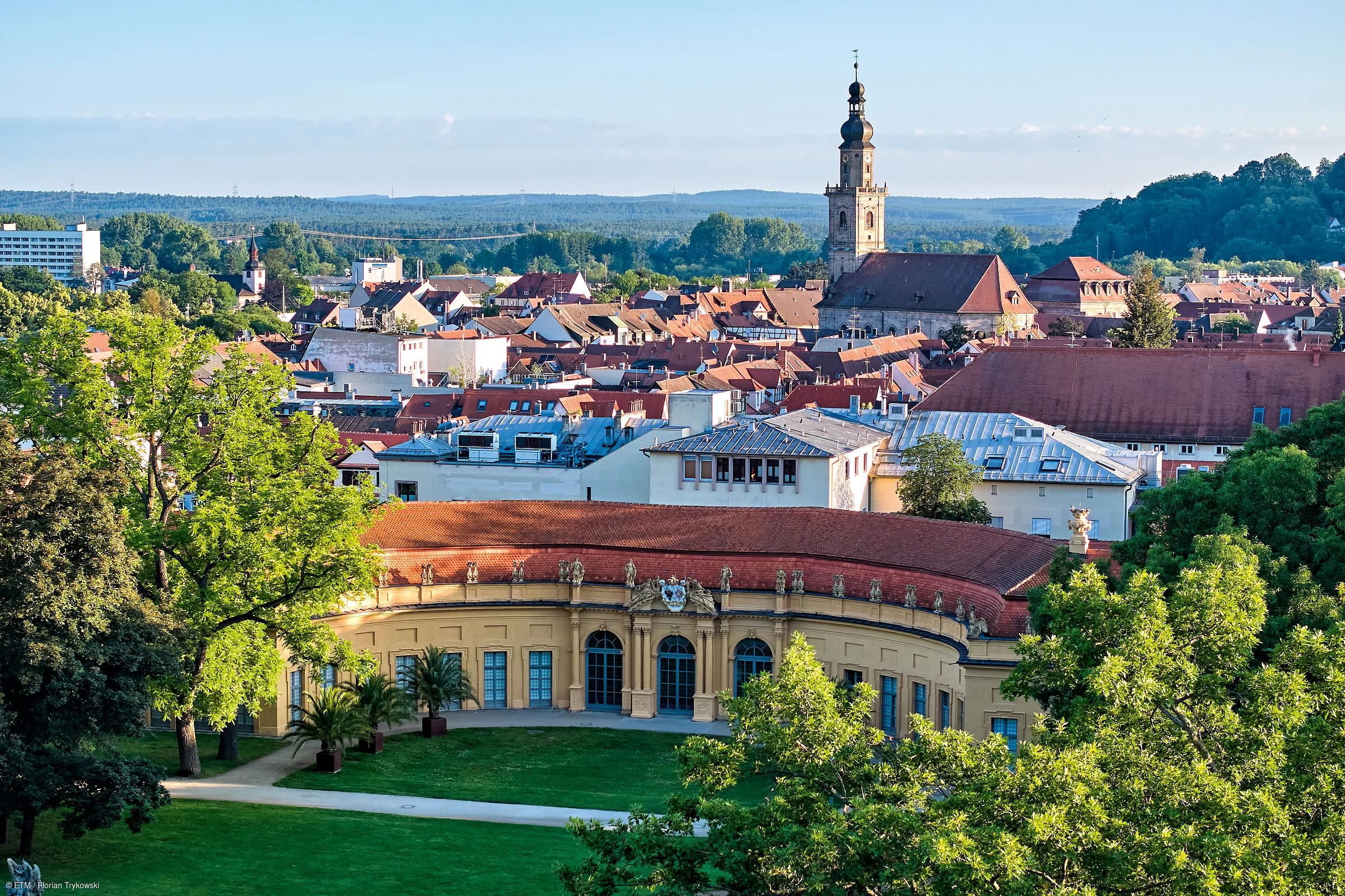 Stadtansicht mit historischem Gebäude, Kirche mit Turm und grünen Bäumen im Vordergrund bei Tageslicht.
