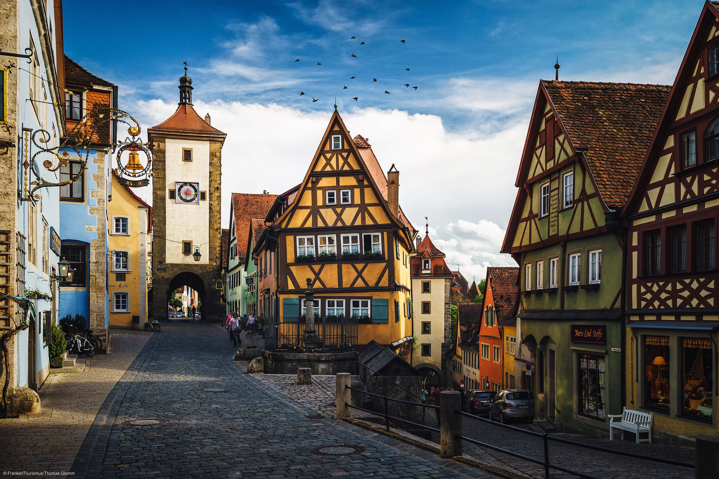 Altstadt mit Fachwerkhäusern, Kopfsteinpflasterstraße und einem Turm mit Uhr unter blauem Himmel mit Vögeln