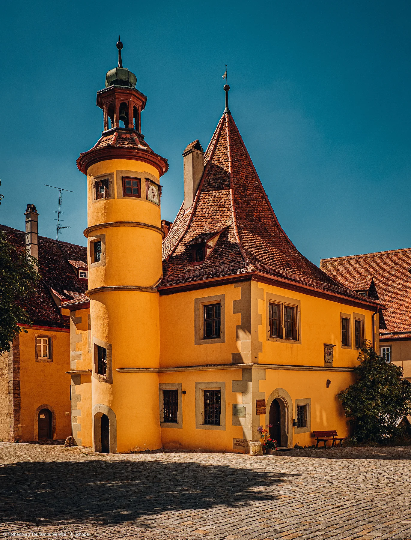 Gelbes historisches Gebäude mit Turm und spitzem Dach bei sonnigem Himmel auf gepflastertem Platz.