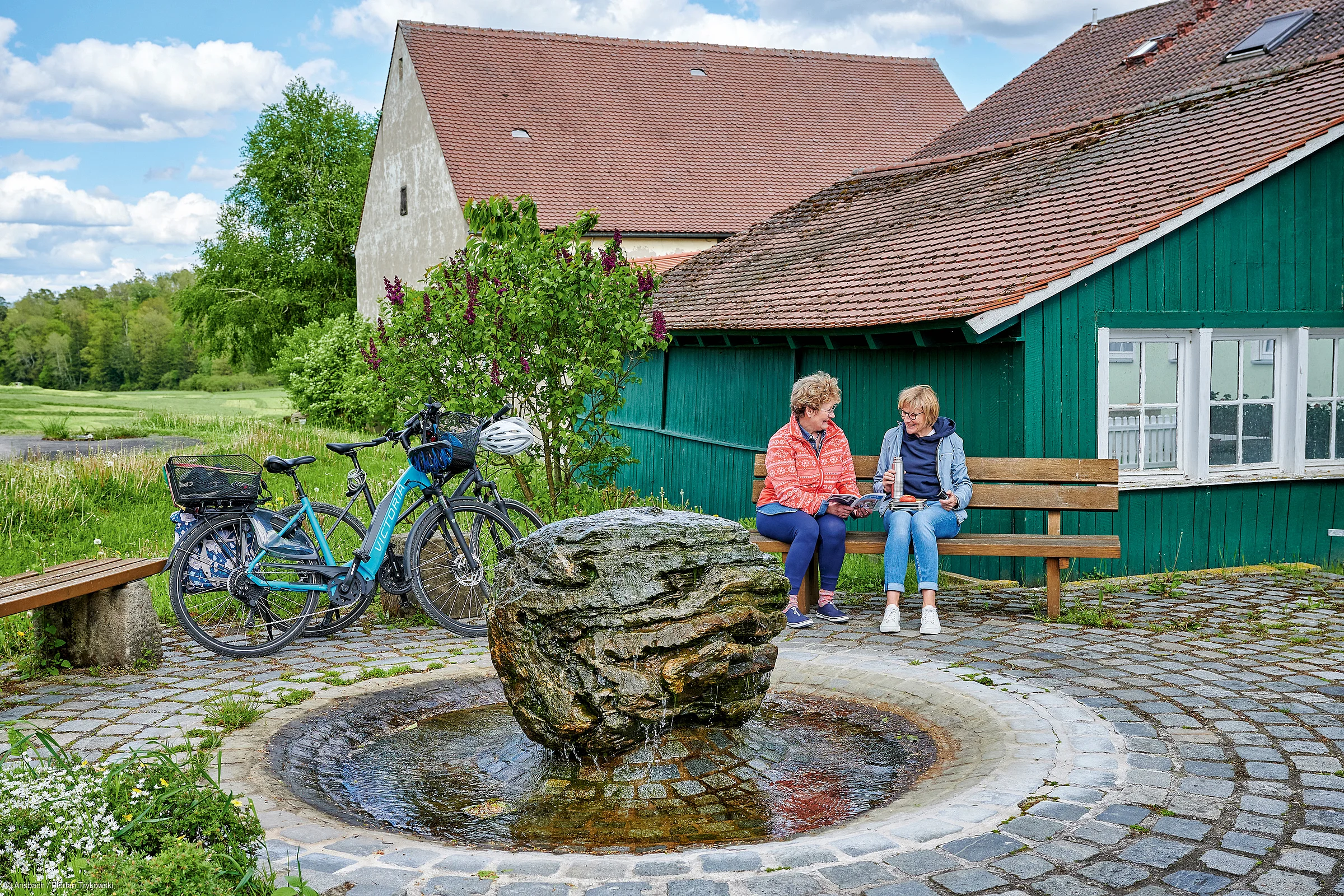 Zwei Frauen sitzen auf einer Bank neben einem Brunnen mit großem Stein, Fahrräder stehen daneben, grüne Holzhütte im Hintergrund.