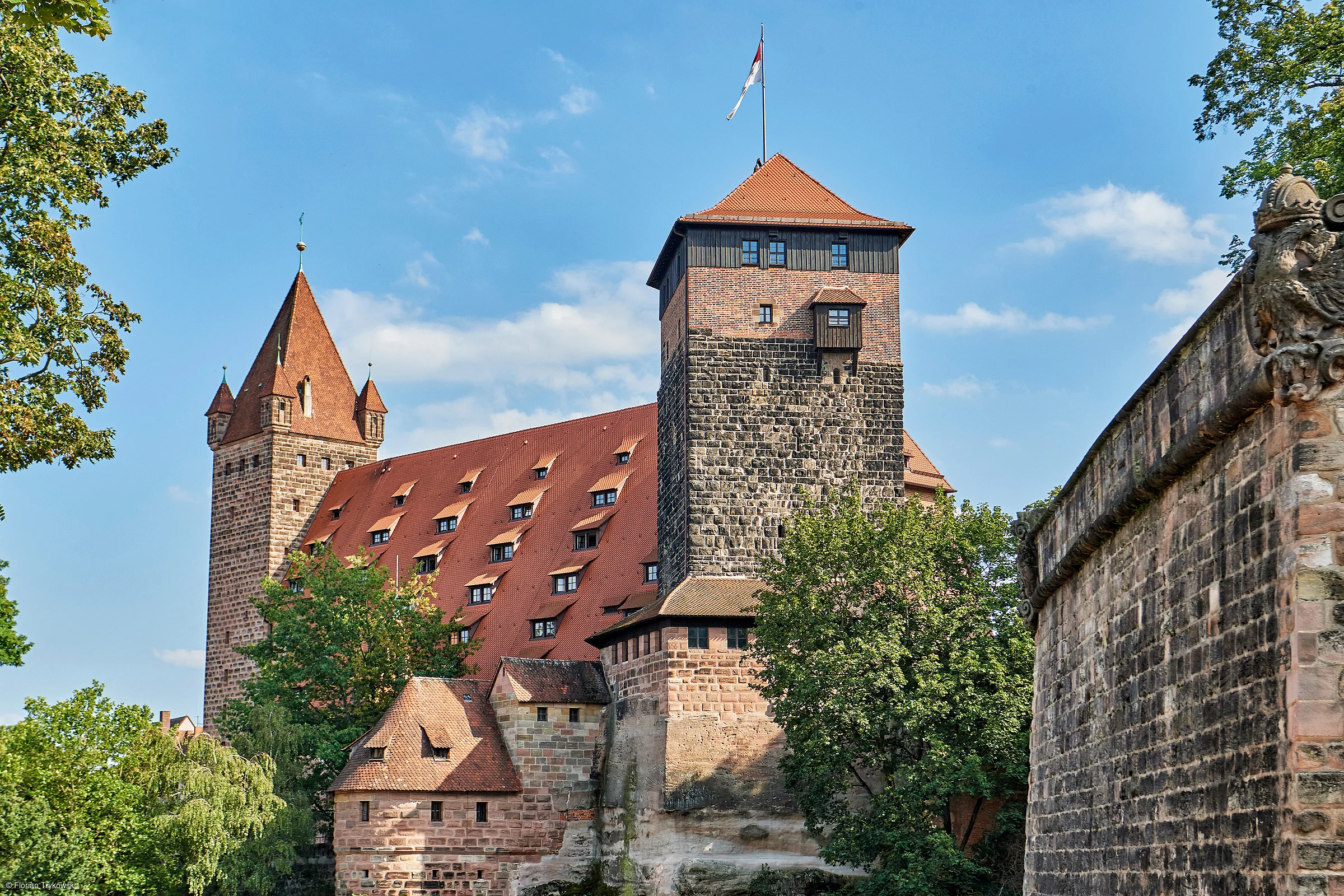 Burg mit roten Dächern und Türmen, umgeben von Bäumen und einer Steinmauer bei blauem Himmel.