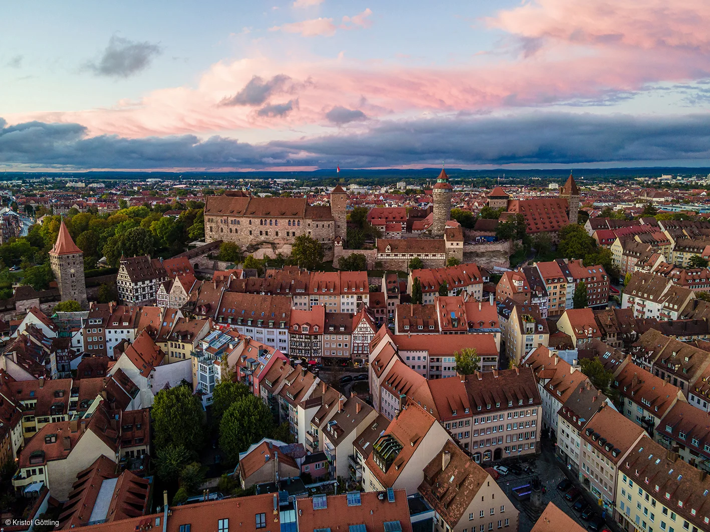Luftaufnahme einer Stadt mit roten Dächern und historischen Türmen bei Sonnenuntergang und bewölktem Himmel.