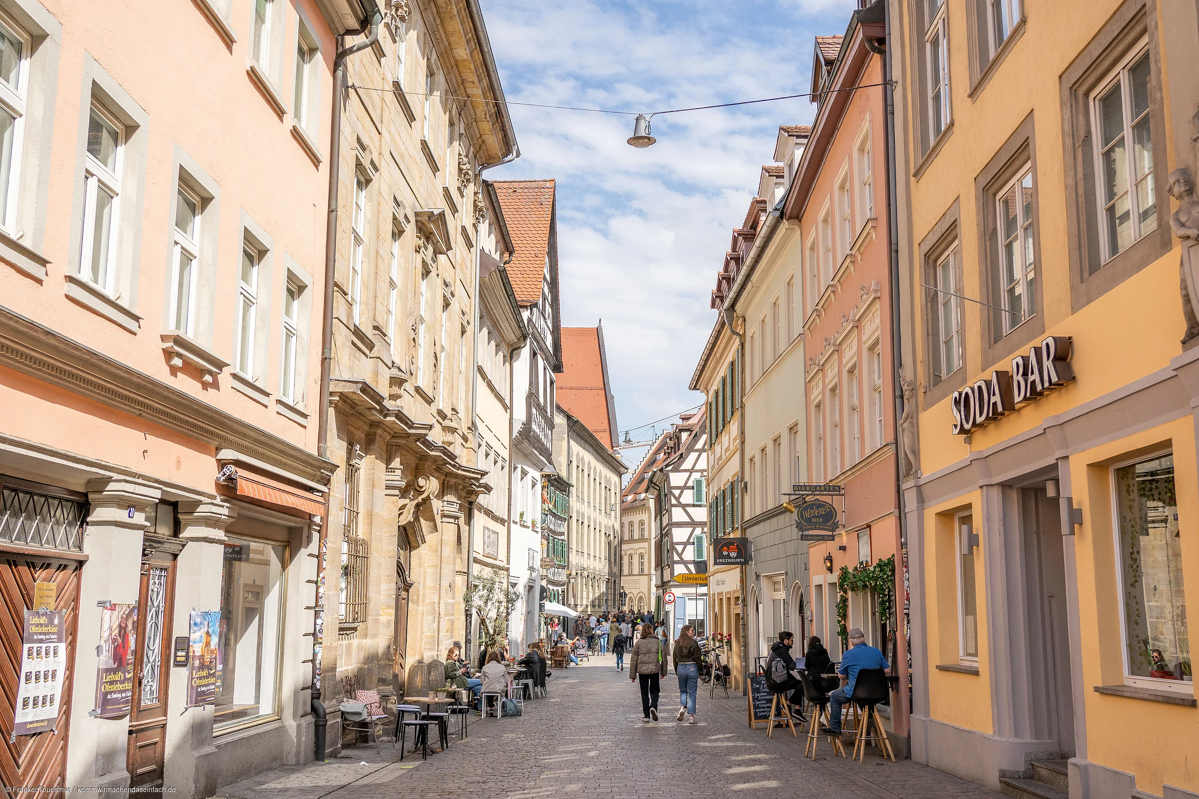 Fußgängerzone mit Cafés, Menschen und historischen Gebäuden unter blauem Himmel mit Wolken.