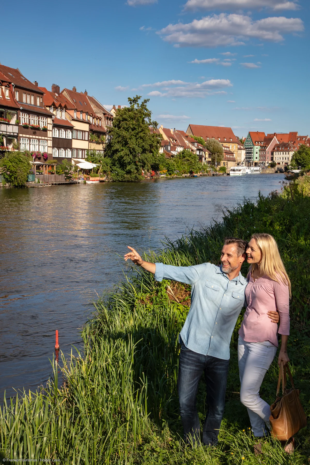 Paar steht am Flussufer, Mann zeigt auf etwas, im Hintergrund Häuser und Boote bei sonnigem Wetter.