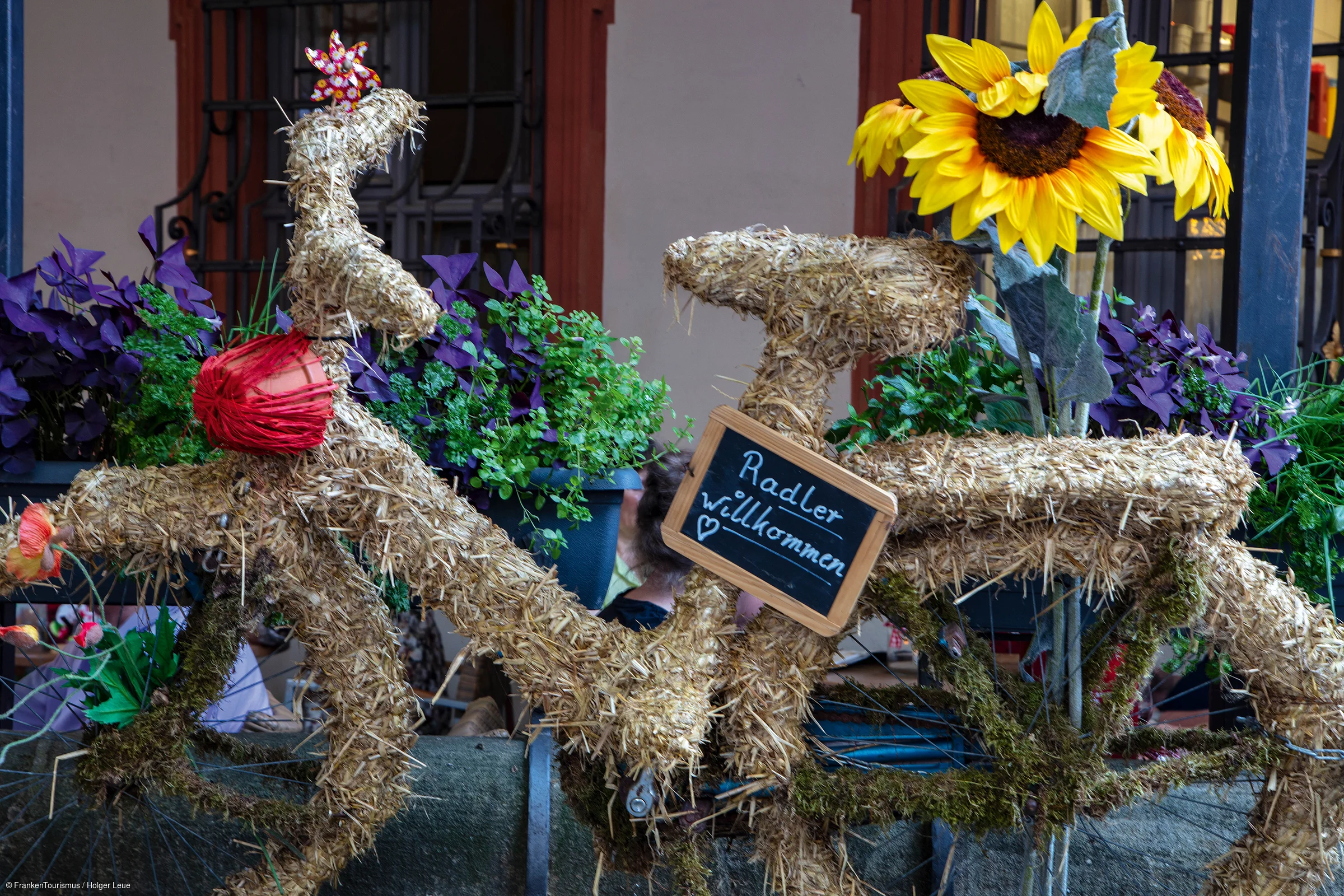 Fahrrad komplett mit Stroh bedeckt, mit Sonnenblumen und Schild „Radler willkommen“ am Rahmen.