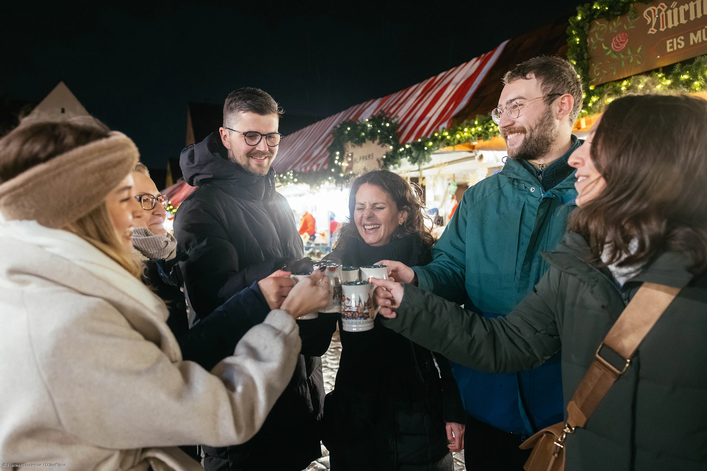 Sechs Personen stoßen mit Tassen bei einem beleuchteten Weihnachtsmarktstand bei Nacht an.