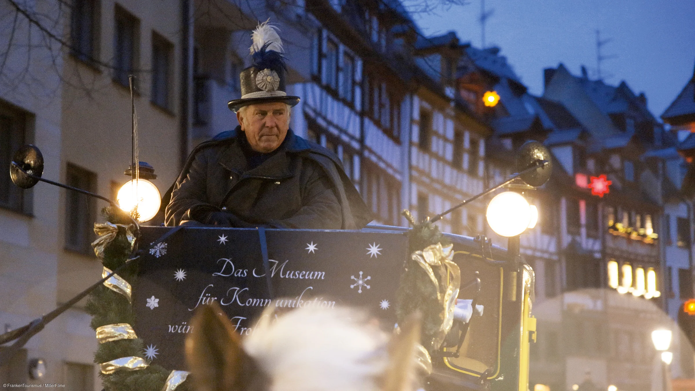 Mann in Postuniform fährt abends in geschmückter Kutsche durch eine Altstadtstraße mit Fachwerkhäusern.