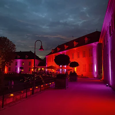 Abendaufnahme eines beleuchteten Gebäudes mit roten und violetten Lichtern und Menschen auf der Terrasse.