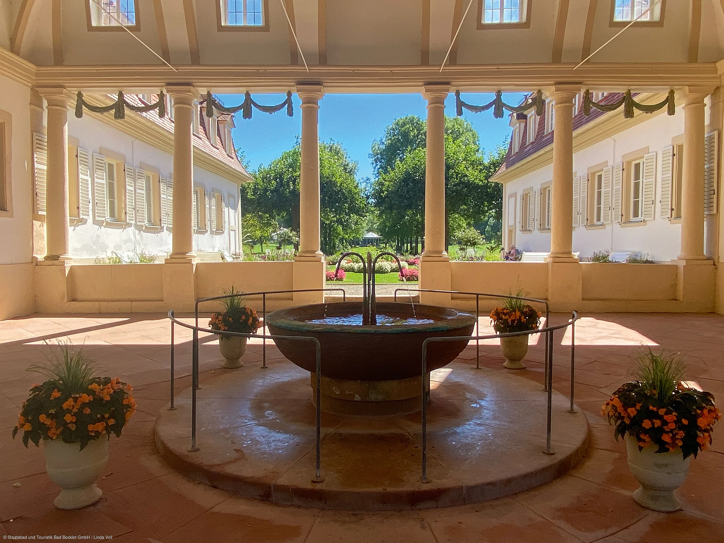 Innenhof mit Springbrunnen, Säulen und Blumendekoration vor einem Garten mit Bäumen und blauem Himmel