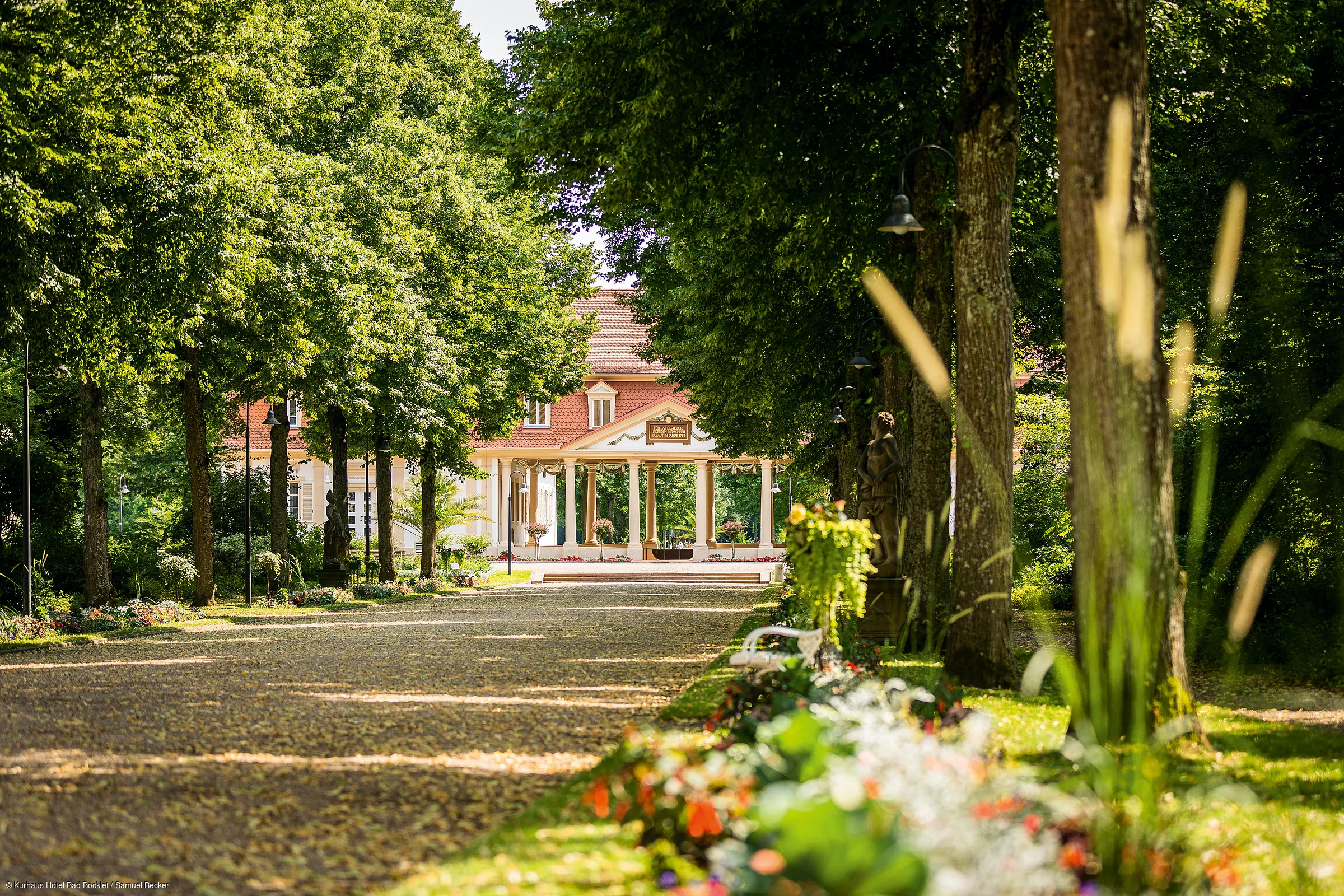 Baumallee mit Blumenrabatten und Sitzbank führt zu einem historischen Gebäude mit Säulenvorbau.