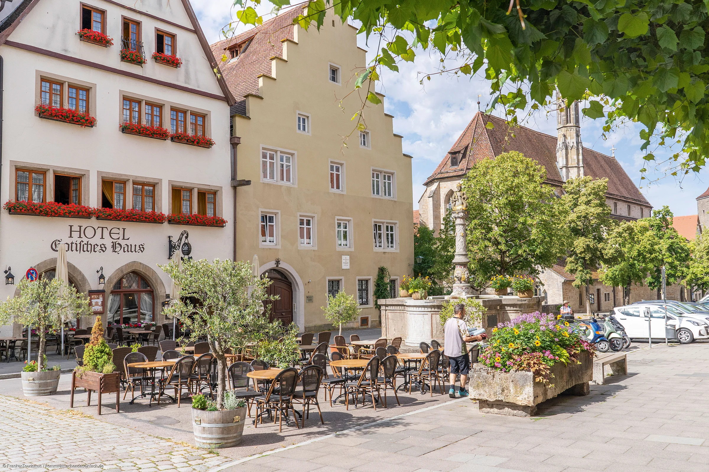 Außenbereich eines Hotels mit Tischen, Stühlen, Blumen und historischen Gebäuden im Hintergrund.