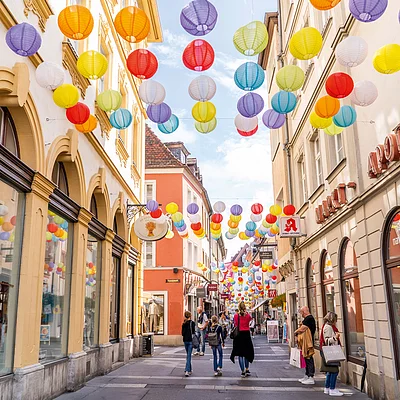 Fußgängerzone mit bunten Lampions über der Straße und Menschen, umgeben von Geschäften.