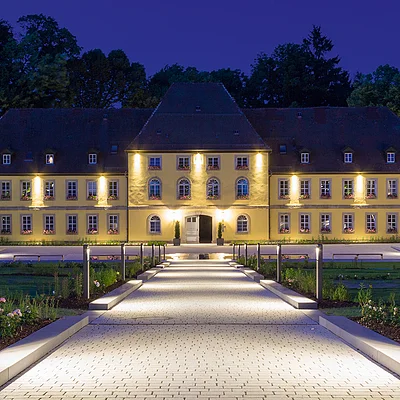 Beleuchteter Weg führt zu großem, gelbem Gebäude mit vielen Fenstern und dunklem Dach bei Nacht.