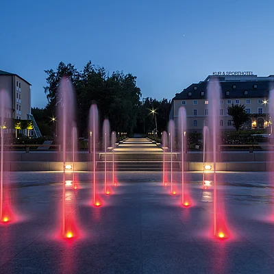 beleuchtete rote Wasserfontänen vor einem Gebäude bei Abenddämmerung mit blauem Himmel