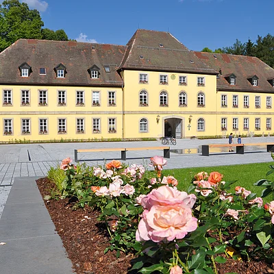 Gelbes mehrstöckiges Gebäude mit vielen Fenstern, davor Rosenbeet und gepflasterter Platz mit Bänken und Brunnen.