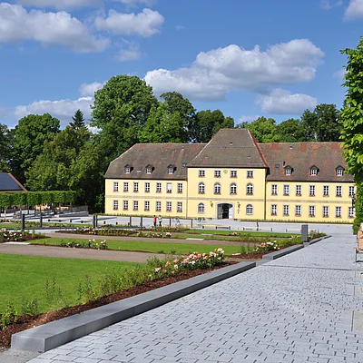Gelbes historisches Gebäude mit großem Garten, gepflastertem Weg und Bäumen unter blauem Himmel mit Wolken.