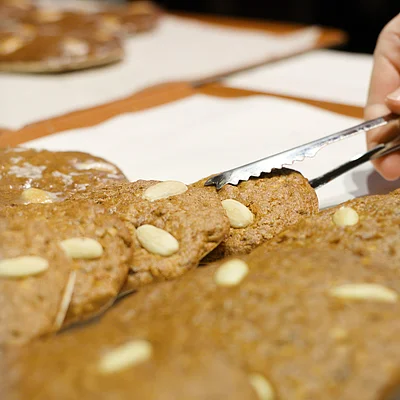 Hand mit Zange nimmt einen Lebkuchen mit Mandeln aus einer Auslage hinter Glas.