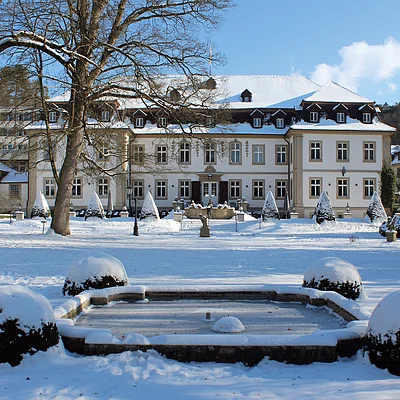 Schneebedeckter Garten mit Brunnen vor einem großen historischen Gebäude und Kirche bei klarem Himmel.