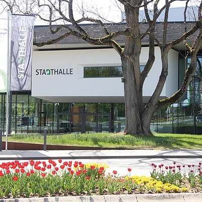 Eingang der Stadthalle mit Fahnen, Baum und Blumenbeeten im Vordergrund bei Tageslicht.
