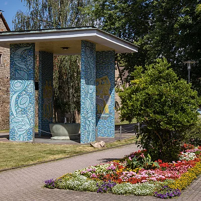Pavillon mit blauen Mosaik-Säulen und Brunnen neben einem Blumenbeet und einem gepflasterten Weg bei Sonnenschein