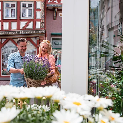Paar betrachtet einen Korb mit Lavendel vor einem Fachwerkhaus und einem Schaufenster mit Pflanzen.