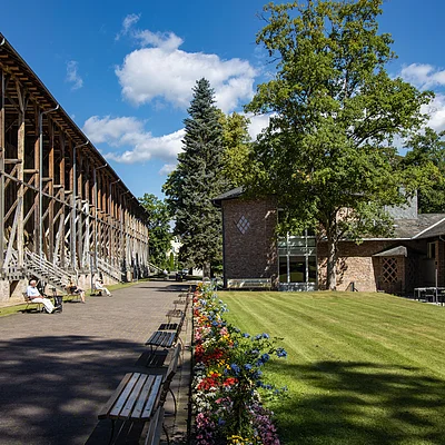 Außenbereich mit Holzkonstruktion, Sitzbänken, Blumenbeet und Gebäuden unter blauem Himmel mit Wolken.
