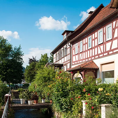 Fachwerkhaus mit Garten und Blumen neben einem Bach unter blauem Himmel mit wenigen Wolken.