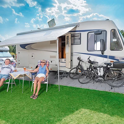 Älteres Paar sitzt unter Markise vor Wohnmobil mit zwei Fahrrädern auf grünem Rasen bei sonnigem Himmel.