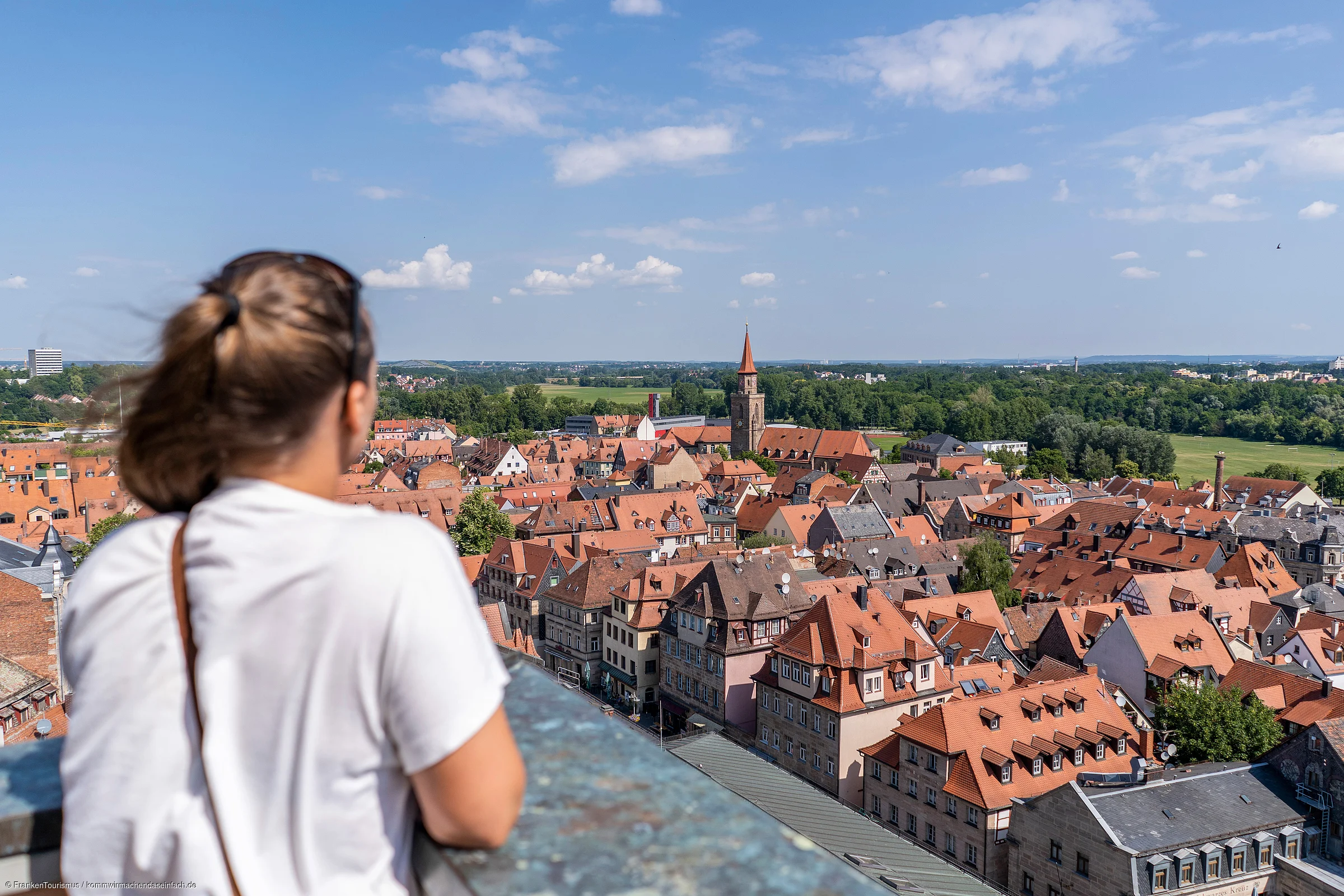 Frau von hinten blickt auf Stadt mit roten Dächern und Kirchturm bei blauem Himmel