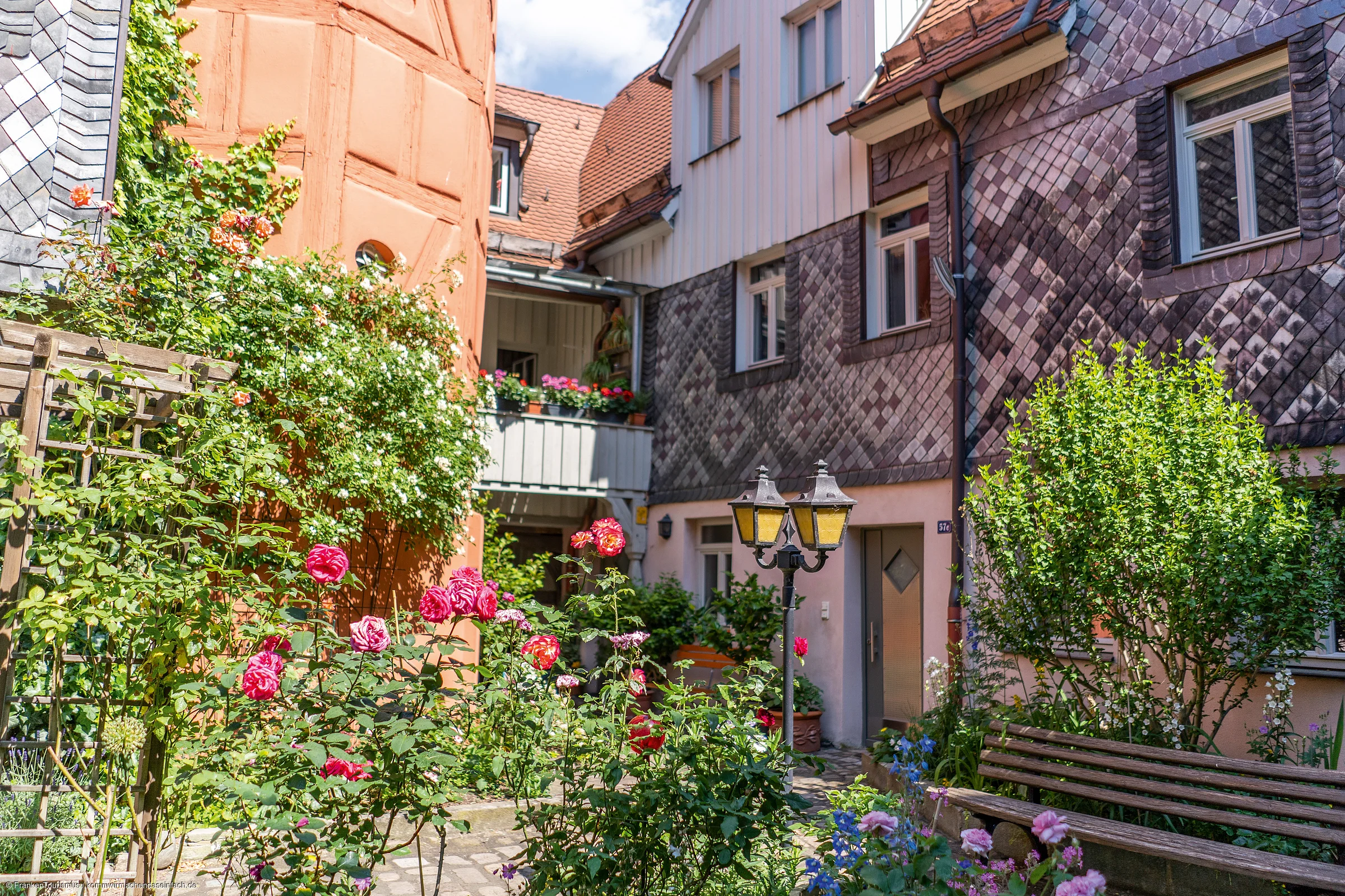 Innenhof mit Rosen, Büschen, einer Bank und historischen Fachwerkhäusern bei Sonnenschein
