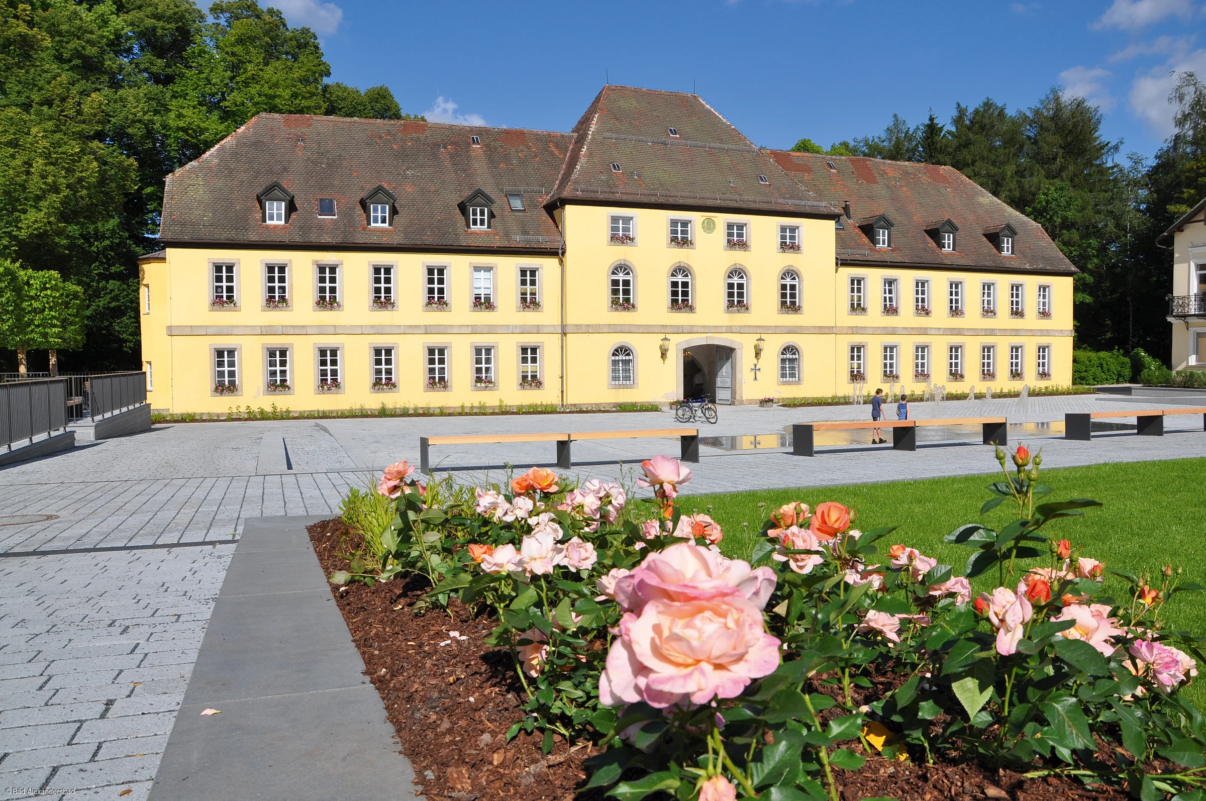 Gelbes mehrstöckiges Gebäude mit vielen Fenstern, davor Rosenbeet und gepflasterter Platz mit Bänken und Brunnen.
