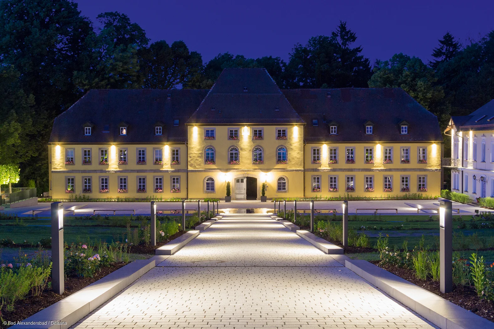 Beleuchteter Weg führt zu großem, gelbem Gebäude mit vielen Fenstern und dunklem Dach bei Nacht.